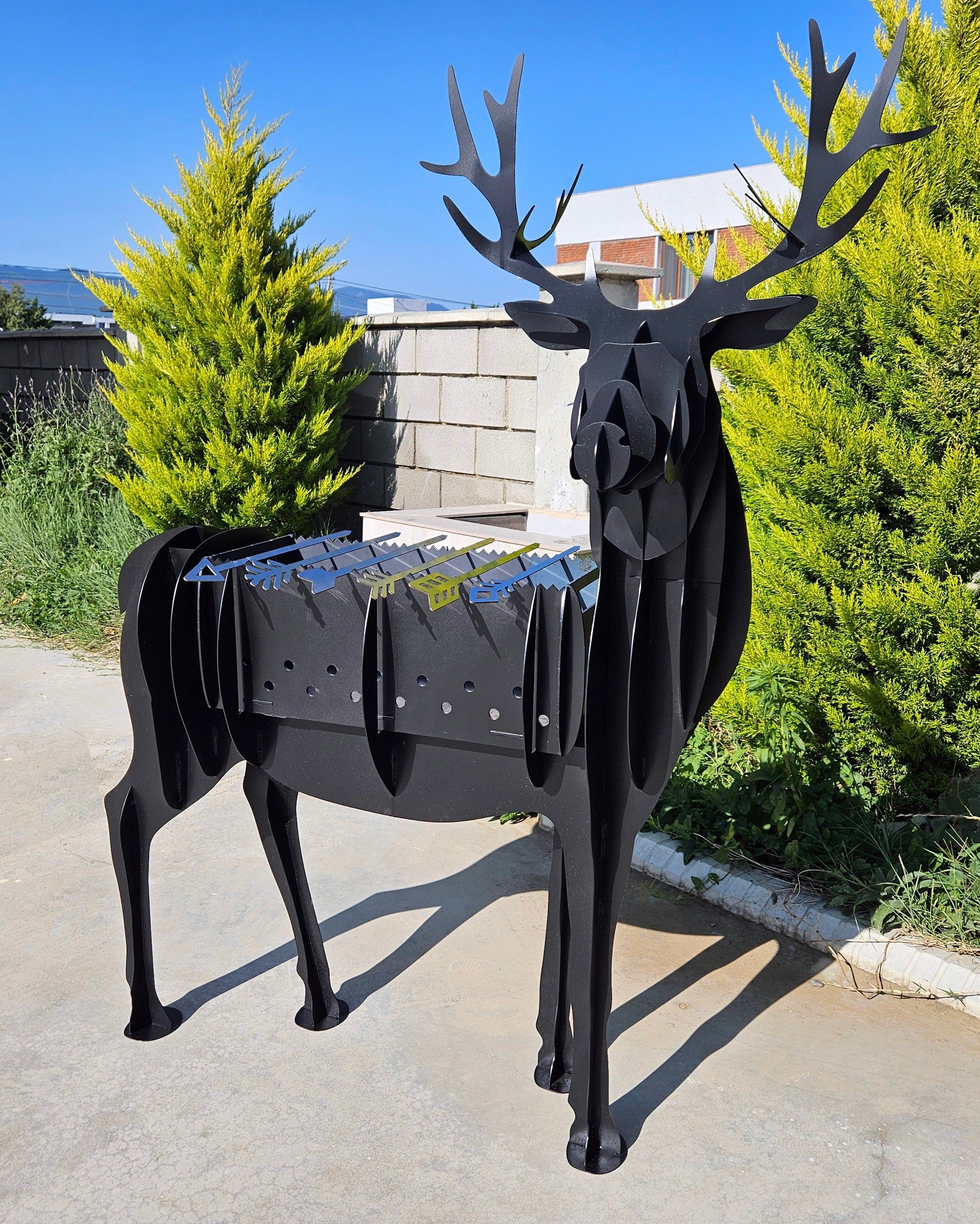 Black metal deer-shaped grill in an outdoor setting with greenery and a clear sky.